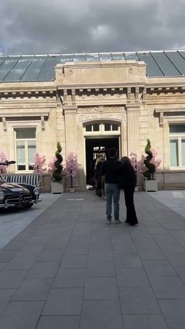Here is a brief description of the video:
The video shows the exterior of a grand building with a glass-paneled roof and ornate stone facade. The building's entrance is flanked by potted pink trees and twisted shrubs. A black and white striped vintage car is parked to the left. In the foreground, two people stand on a tiled terrace. Emerging from the building's entrance, a woman in a fur coat and shorts walks with a man in a suit. Another woman is also seen leaving the building. The sky above is cloudy.