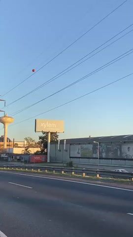 The video shows a street view from a moving vehicle, revealing an industrial area under a clear blue sky. On the left side of the road, a tall utility pole supports a network of power lines. In the distance, a white water tower stands next to buildings, displaying flags. A large billboard advertising “Xylem” is visible along with other advertisements. The road appears to be paved, with some guardrails and fencing separating it from adjacent structures. A solitary red ball hangs suspended from one of the power lines, attracting attention against the skyline. A car passes on the right side of the road.