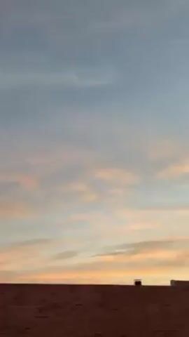 The video pans up from a brick building to reveal a sunset sky with a mixture of blue, orange, and pink clouds. The clouds are spread out across the sky, with the orange hue concentrated near the horizon. As the camera pans up, residential buildings appear in the distance, with a mountain range visible beyond them. The overall scene is peaceful and serene.