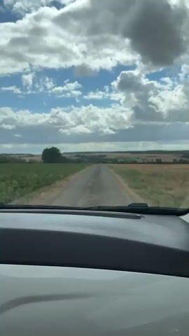 The video captures a first-person perspective from inside a vehicle, looking out through the windshield at a rural landscape. The vehicle is on a paved road that stretches into the distance, flanked by fields of green crops on the left and dry, golden fields on the right. The sky is filled with dramatic, puffy white clouds against a blue backdrop. As the vehicle moves forward, the road leads towards a gentle incline, beyond which a single, round hay bale is visible in the right-hand field. The horizon shows a varied landscape of trees and more fields, with some distant structures or trees suggesting a rural settlement. The dashboard of the vehicle is visible at the bottom of the frame.
