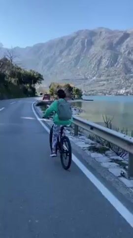 A person is riding a bicycle on a paved road. The person is wearing a green jacket, patterned pants, pink sneakers, and a dark cap. They also have a grey backpack. The road has a white line marking the edge and a metal guardrail on the right. To the right of the road is water, and behind the water are large mountains. A red car is visible in the distance on the road ahead. The sky is blue. The camera follows the cyclist.