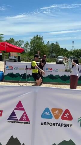 A video of a medal ceremony shows a man in a yellow shirt with a hydration pack receiving a medal from a woman on the left and shaking hands with a man on the right. Another man in a black shirt speaks into a microphone on the left. A white banner with logos and text is in the foreground. A red tent and various people are in the background, with a bright blue sky above.