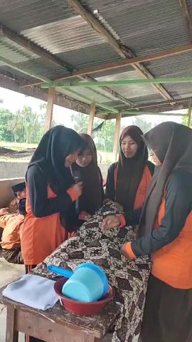 Four young women wearing hijabs and orange and black uniforms are gathered around a table. One woman on the far left is holding a microphone and appears to be speaking. Another woman in the center is holding a blue plastic ladle and a patterned cloth. The third woman from the left is watching the woman with the ladle. The fourth woman on the far right is also watching the cloth. A red bowl and a white cloth are on the table. In the background, another person wearing an orange and brown uniform is seated. The scene appears to be outdoors under a corrugated metal roof supported by wooden beams.