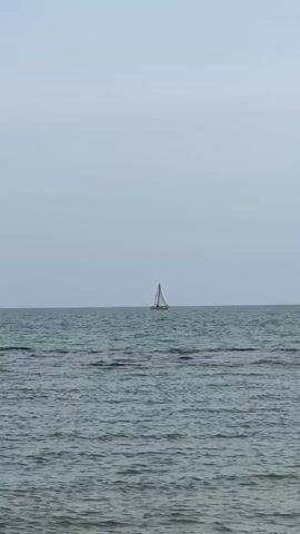 The video shows a sailboat on a calm sea under a cloudy sky. The boat, which appears to be a small sailboat, is in the distance and is the focal point of the shot. The sea is relatively still, with small waves. The sky is overcast and a uniform light gray color.