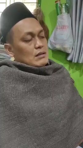 A man wearing a black taqiyah and a grey shawl is shown in a close-up shot. His eyes are closed, and he appears to be in prayer or meditation. The background is slightly out of focus, but shows a green wall, a window with white bars, and some items hanging on the wall. The lighting is dim.