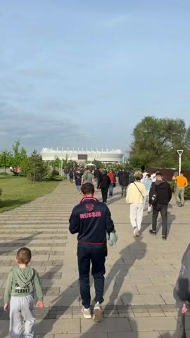 A crowd of people is walking on a paved walkway toward a large stadium under a partly cloudy sky. The camera follows behind the crowd, focusing on a man in a navy blue tracksuit with the word "RUSSIA" and a Russian coat of arms on the back. A young boy is walking next to him. Other people in the crowd are dressed in casual clothing. The walkway is flanked by grass and trees. The stadium is white with a large screen visible above the entrance.