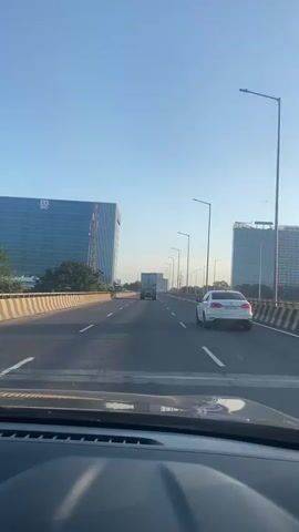This is a first-person perspective video, likely from inside a vehicle, traveling on a multi-lane highway. The sky is clear and blue. On the left side of the highway are two large, modern glass-fronted buildings. On the right side, there are more similar buildings and trees. The highway has yellow and black striped barriers along the sides and white lane markings. There are streetlights visible along the highway. Several vehicles are on the road: a white car is in the right lane ahead, and a truck is in the left lane further ahead. The lower portion of the video shows the dashboard of the vehicle from which the video is being recorded. The audio appears to be music with Tamil lyrics.