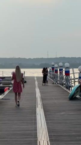 A blonde woman in a red polka-dot dress walks away from the camera on a wooden pier towards a body of water. Further down the pier, a couple embraces. The sun is setting, casting a warm glow on the scene. Several boats are docked along the side of the pier. The sky is cloudy with streaks of orange and yellow.