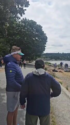 The video begins by focusing on two people walking away from the camera on a paved path. The person in the foreground on the left is wearing a blue jacket, a white visor, and shorts. The person in the foreground on the right is wearing a dark blue jacket and a dark baseball cap. Behind them, another person in a white jacket and pink pants is walking towards the camera. The path is bordered by a grassy area on the left and a rocky, debris-strewn area on the right.

The camera then pans to the right, revealing a shoreline with a calm body of water and a distant tree-lined coast. There are several people on the beach, some appearing to be engaged in some form of cleanup or gathering debris. Among the debris are numerous pieces of driftwood and logs. In the distance, a ferry can be seen on the water. The sky is overcast with clouds. The video continues to pan across the beach and the water, showing more driftwood and the overall landscape. The camera then appears to move further along the path, looking out towards the water and the distant shore, with more driftwood scattered along the beach. The video concludes with a wide shot of the water and the cloudy sky.