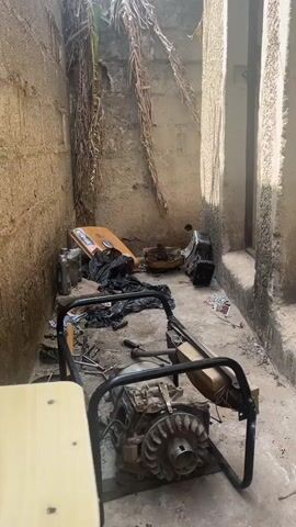 The video shows a cluttered outdoor space.  The walls are made of cinder blocks and concrete, and there are dead leaves and vines hanging from the top.  A generator frame with the engine inside sits in the middle of the area.  There are tools scattered around the generator, along with pieces of what appears to be the disassembled generator casing. A wooden stool stands to the left.  A partly open door is visible on the right side of the frame. Debris is scattered on the ground.