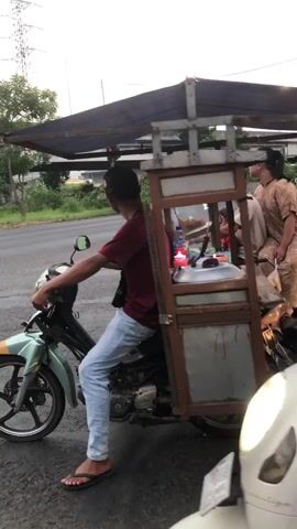 The video shows a man riding a motorcycle with a cart attached to the back. The cart is covered with a tarp and appears to be used for selling food. A woman is sitting in the cart behind the man. The motorcycle is green and white, and the cart is made of wood and metal. The video is taken from the perspective of another motorcycle, which is slightly in front and to the right of the main subject. The environment appears to be a road with some trees and vegetation in the background. The lighting suggests it is either early morning or late afternoon.