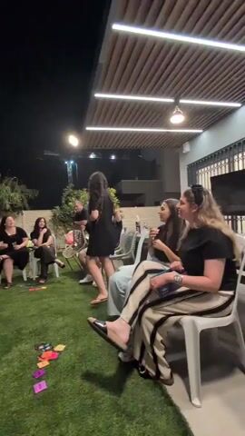 This video shows a group of people sitting on chairs in a grassy backyard at night. There is a table in front of some of the people with food and drinks on it. A woman in a black dress is standing in the middle of the group and appears to be leading an activity or game. The other people are seated and observing her. The environment is outdoors with trees and a fence visible in the background. The lighting suggests it is evening or night.