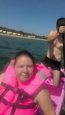 A young woman wearing a pink life jacket smiles at the camera while sitting on a pink paddleboard. A young man wearing black shorts kneels behind her, holding a paddle. They are on the water with a beach and buildings in the background. The water is choppy and the sky is clear and blue.
