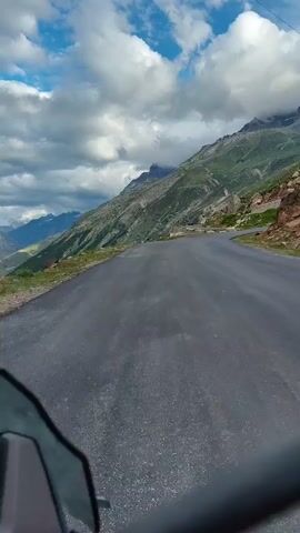 The video shows a first-person perspective of a motorcycle or vehicle driving on a paved road. The road is winding and appears to be going uphill, with a steep mountain slope on the right side of the frame. The mountain is covered in green vegetation and rocks, with some areas of exposed earth. The sky is partly cloudy, with dramatic white clouds against a blue sky. In the distance, more mountains are visible, some with snow-capped peaks. The left side of the frame shows the vehicle's windshield or fairing in the foreground. The overall impression is a scenic drive through a mountainous landscape.