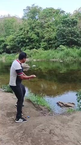 The video shows a man standing on a dirt bank next to a calm river throwing a rock into the water. The man is wearing a white and black t-shirt, black pants, and black and white sneakers. The river is surrounded by lush green trees and vegetation. The sky is overcast.