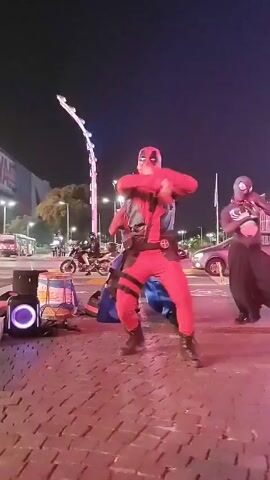 Three people in superhero costumes are dancing on a red brick-paved street at night. The middle figure wears a Deadpool costume, while the two on either side are dressed as Spider-Man variants. They are performing in a synchronized, energetic manner. Behind them are streetlights, buildings, parked cars, and a parked motorcycle. The sky is dark. The scene appears to be a street performance in an urban setting.