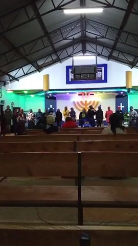 A medium shot from behind wooden pews shows people on a brightly lit stage in what appears to be a church or large meeting space. The stage is decorated with green, blue, and purple lights and what appears to be a menorah and crosses. Many individuals are standing, and some appear to be singing or performing. A few people are seated in the pews. The roof is a tall, industrial-style structure.