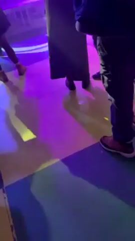 People are gathered in an indoor venue, possibly a club or event space. There is a DJ performing on stage. The lighting is dynamic, featuring a mix of white and purple spotlights. Banners with logos or advertisements can be seen in the background. The crowd appears to be enjoying the music and atmosphere.