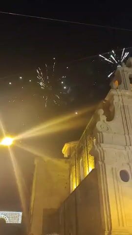 This is a video of fireworks being set off at night in front of a church. The church is lit up with yellow lights. There are many people gathered in front of the church watching the fireworks. The fireworks are colorful and bright, and they are exploding in the sky. Some of the fireworks are leaving trails of light behind them. The video is shaky, and it is difficult to see some of the details.