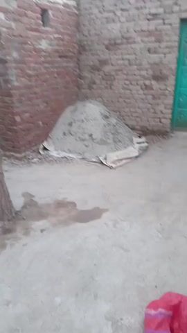 The video shows an outdoor scene with a pile of dirt and a brick wall. A tree is visible on the left side of the frame. In the background, there is a green door. In the center of the frame, a person wearing a light blue outfit is visible, adjusting their hair and holding a stick. The ground appears to be dirt and concrete, with some puddles of water. A red and pink bag is on the ground on the right side of the frame. The video is shaky and the focus is not sharp.