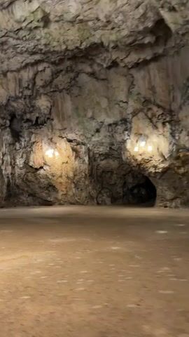 A low-angle shot within a cave reveals a vast, rugged ceiling and walls adorned with numerous stalactites and stalagmites. Several circular light fixtures illuminate the cave's natural formations. A yellow barrier is visible on the left. The camera pans right, showing a person with a black cap and a blue jacket holding a smartphone to record the cave. The camera continues to pan right, revealing the backs of multiple people seated in yellow, boat-like vehicles. The cave floor is flat and sandy.