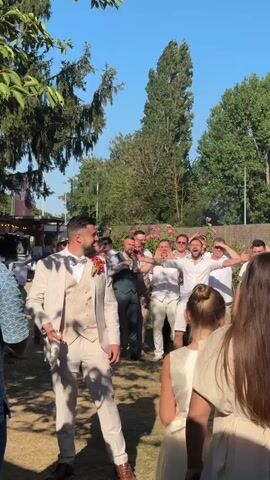 A man in a light-colored suit stands in a grassy outdoor area, holding a rolled-up paper and smiling. He is surrounded by other men, some of whom are in white shirts and pants. The man in the suit turns to his right and waves, and the other men cheer and wave back. In the foreground, two women in light-colored dresses are visible from behind. The background features trees and a clear blue sky.