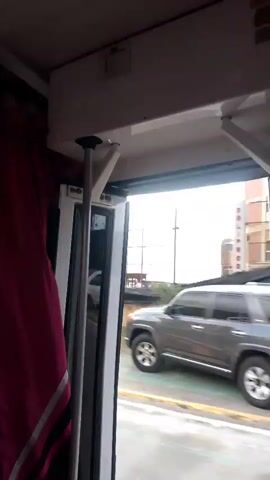 The video is shot from inside a vehicle, likely a bus or train, looking out an open doorway. The perspective is slightly tilted. To the left, a maroon curtain is partially visible. The doorway frames a street scene. A silver SUV is moving from right to left across the frame, its tires blurred due to motion. The road appears wet or reflective. In the background, a tall, light-colored building with multiple windows can be seen, and further in the distance, a red structure is partially visible. The sky is overcast. The overall impression is of travel on a city street.