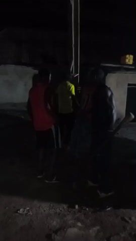 The video shows a group of young men in a dimly lit outdoor environment at night. They appear to be gathered around a wooden pole. One man on the right is holding what looks like a bat or stick. The man in the red shirt in the foreground is facing away from the camera and seems to be interacting with the others. The background suggests a rural or semi-urban setting with simple structures and debris on the ground. The lighting is poor, making it difficult to discern fine details.