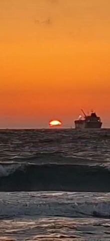 The video captures a serene ocean sunset. The sun, a small, bright orange orb, is partially submerged below the horizon, casting a warm glow across the sky. The water is a deep, dark blue, with gentle waves rolling towards the shore. In the distance, a large cargo ship is silhouetted against the vibrant sky, its mast and cranes visible. The overall scene evokes a sense of peace and tranquility.