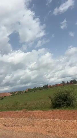The video shows a landscape from a moving vehicle. The foreground consists of a rough, reddish-brown surface, possibly a dirt road or unpaved shoulder. Beyond this, a vibrant green field stretches out, appearing to be a grassy meadow or pasture. In the middle ground and background, rolling hills are visible. These hills are characterized by large mounds of reddish-orange earth, suggesting bare soil or exposed mineral deposits. Patches of green vegetation, including trees and shrubs, are scattered across the hillsides and in the foreground. The sky above is a mix of blue and white clouds, indicating partly cloudy weather. The camera appears to be angled slightly upwards, capturing the expanse of the field and the sky. The overall impression is of a rural or natural environment, possibly a mining or geological area due to the exposed earth.