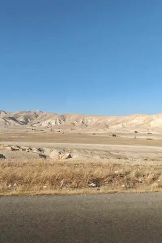This video shows a landscape view from a moving vehicle. The foreground features a patch of dry grass and a road barrier. Beyond that, a barren, arid landscape unfolds with rolling hills and mountains in the background. The sky is a clear, deep blue. Scattered trees and shrubs are visible in the mid-ground. The overall impression is of a desert or semi-arid environment. There are no people or specific actions depicted, other than the implied movement of the vehicle.