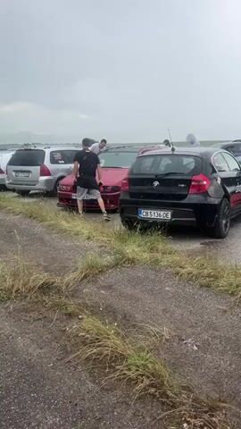 The video shows a gathering of cars in what appears to be a parking area on a rainy day. Several individuals are standing around the vehicles, some of which seem to be modified for performance. The cars include a black BMW 1 Series, a red BMW 5 Series with custom headlights and aftermarket body kit, and a grey Subaru. Engine revving noises can be heard in the background. The environment looks like a gravel lot with patches of weeds growing through the surface. The sky is overcast, suggesting persistent rain.