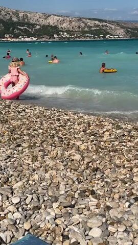 A child wearing a pink bathing suit and a hat is sitting on a pink inflatable donut float on a pebble beach. The child is facing away from the camera, looking out at the ocean. Several other people are in the water, some swimming, others floating on yellow inflatables. The water is a bright turquoise color, and there are waves. In the background, there are rocky hills with some buildings on them. The sky is blue and clear.