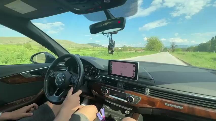 From the interior of an Audi A5, a person is driving on a narrow asphalt road. The driver is wearing a short-sleeved shirt and is holding the steering wheel with both hands. The car’s infotainment system displays a map.  Outside the car, the sky is blue with fluffy white clouds. The road is flanked by grassy fields and occasional trees. In the distance, there are low-lying mountains. The overall scene suggests a drive through a rural, possibly agricultural area on a sunny day. The car has wooden trim.