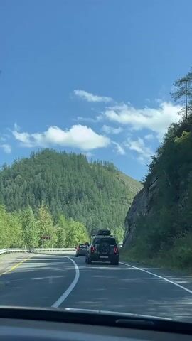 The video shows a view from inside a car driving on a paved road. The road curves to the left, with white lines marking the lanes. On the left side of the road, there is a green, tree-covered mountain. On the right side of the road, there is a rocky cliff face. Several cars are visible on the road ahead, including a black SUV with luggage on its roof, a white van, and other passenger vehicles. A blue and white truck with "OZON" written on it is also visible in the distance. The sky is blue with scattered white clouds. The overall impression is that of a scenic drive through a mountainous, forested area.