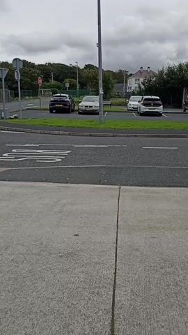 A black car is driving down a street. There are other cars parked on the side of the road. There is a sidewalk in the foreground and a sky with clouds in the background.