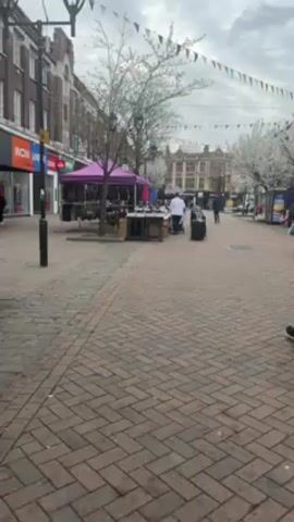 The video shows a brick-paved pedestrian zone in a town. Several market stalls are set up along the street, one with a purple canopy. People are walking around, some pulling luggage. There are buildings on either side of the street, and decorative pennants are strung overhead. Some trees are in bloom with white flowers. The sky is overcast.