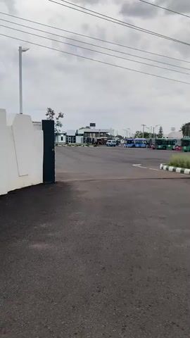 The video opens with a view of an empty asphalt lot, likely a parking area. A white concrete wall and a black gate are visible on the left, suggesting an entrance. Beyond the gate, a wider view of the lot reveals several parked buses of various colors, including blue, green, and pink. Some smaller vehicles, possibly vans or minibuses, are also present. In the background, a building with a white facade and dark roof is visible, with a few streetlights scattered across the scene. The sky is overcast with gray clouds. The video pans slightly to the right, offering a clearer view of the parked buses.