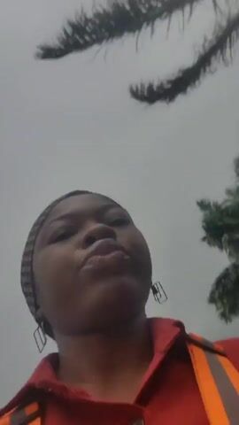 A woman in a red shirt with reflective orange and white stripes is taking a selfie video. She is wearing a patterned headband. Behind her, a silver SUV is parked in a lot with yellow lane markings. The background shows some greenery and industrial buildings. The video is vertically oriented.