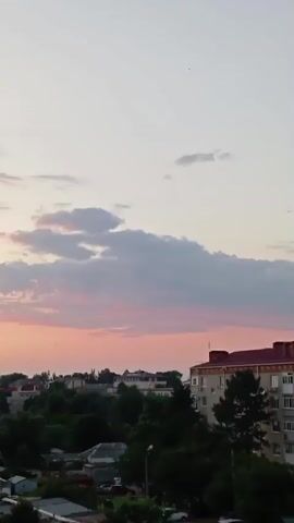 This is a vertical video of a sunset over a town. The sky is filled with soft pink and orange clouds, transitioning to a pale blue at the top. Several small birds are visible flying in the sky. Below the sky, the landscape features a collection of buildings, including what appears to be residential apartment buildings and smaller structures that might be garages or workshops. Lush green trees are interspersed throughout the scene, with some closer to the foreground and others forming a line between the buildings and the sky. The overall impression is a peaceful, late afternoon or early evening in a semi-urban environment. There is no visible text in the video.