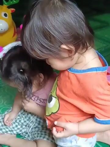 Two young children are sitting on a green tiled floor. The child on the right, wearing an orange t-shirt and a diaper, is holding the t-shirt of the child on the left, who is wearing a pink shirt and a plaid skirt. The child on the left is looking down and appears to be picking at something near their mouth. In the background, a yellow and red stuffed reindeer toy is visible. The lighting suggests an indoor setting.