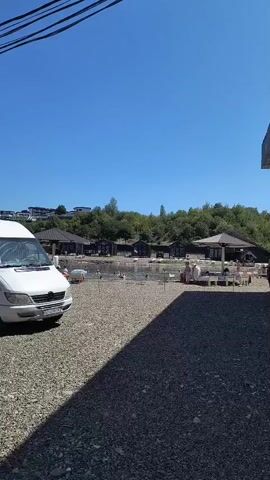 A white van is parked on a gravel lot. Beyond the lot, there is a body of water with people swimming. Several small structures with dark roofs are visible along the edge of the water. In the background, a wooded hill rises under a clear blue sky. Some people are seated at tables under umbrellas on the gravel lot.