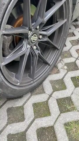 This is a close-up, medium shot of a dark gray Nissan wheel on a car. The car is parked on an interlocking paver surface, which is common for driveways or parking areas. Some patches of green grass are visible between the pavers, suggesting an outdoor setting. The wheel has a multi-spoke design and a visible Nissan logo in the center. The tire is dark and shows some wear. The brake disc behind the wheel appears rusty. The lighting suggests it is daytime, and the weather appears to be overcast or dull, as there are no harsh shadows. The image is static and focuses solely on the wheel and its immediate surroundings. No people are visible. The visible text on the tire reads "BRIDGESTONE" and "CBN40208."