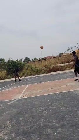 Two men play basketball on an outdoor court under a cloudy sky. One man in a black t-shirt and gray shorts dribbles the basketball, then shoots a jump shot. The basketball flies towards the basket, which is surrounded by tall grass and a chain-link fence. The man in the black tank top and black shorts watches the shot.