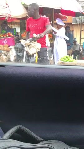 This media file captures a bustling market scene in Aketu-Oja, Nigeria. The focus is on a man on a motorcycle, wearing a red t-shirt and dark pants, looking to his right. He appears to be interacting with someone off-camera or preparing to move.

In the background, several other individuals are visible. A woman in a white outfit stands to the right of the motorcyclist, holding a white object. Further back, a man in a blue shirt is seated. Another person in a brown robe walks past, holding hands with a young child wearing a patterned dress.

The setting is an outdoor market with various stalls and goods on display. Piles of pineapples are visible on the left, along with boxes of fruit, and a display of green produce, possibly limes or lemons, in the foreground. Umbrellas provide shade, suggesting it is daytime. The presence of water droplets on the window in the foreground indicates that it might be raining or has recently rained. The overall atmosphere appears lively and active, typical of a busy marketplace. No specific text is legible in the file.