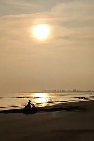 The video shows a beach at sunrise. The sun is a bright circle in the sky, casting a golden glow on the water. A person on an ATV drives along the beach, then the ATV leaves the scene. The beach is sandy and wet, with gentle waves lapping at the shore. In the distance, there is a hazy land mass with trees. The sky is pale yellow.