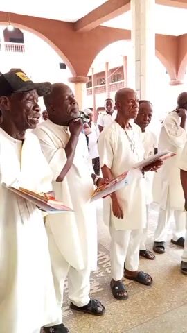 A group of elderly African men are standing in a row, singing from what appears to be song sheets. The men are dressed in white clothing and sandals. One man is wearing a black baseball cap. The scene is taking place in what looks like a covered hallway or courtyard with arches and a multi-story building in the background. Some other people are visible in the background. The men appear to be focused on their singing.
