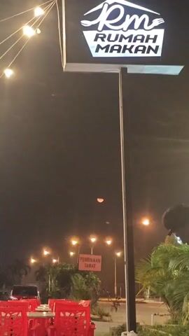 A low-angle, nighttime shot captures a sign for "RM RUMAH MAKAN" illuminated by string lights. Below the sign, a person wearing a dark shirt and a baseball cap is visible, with their back to the camera, looking down. Red tables and chairs are set up in front of what appears to be an outdoor seating area for the restaurant. In the background, streetlights illuminate a road and palm trees. A sign that reads "PEMBINAAN TAMAT" is also visible. The sky is dark with a faint red crescent shape, possibly a reflection or an object in the sky. The lighting suggests a humid or rainy atmosphere due to the haze.