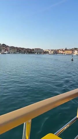 From the perspective of a boat, the video shows a panoramic view of a coastal town across a body of water. The town consists of closely spaced buildings with varied colors, set against a clear blue sky. A prominent bell tower stands out on the left side of the town. The water is calm with gentle ripples, and a yellow railing is visible along the lower edge of the frame, indicating the perspective is from a moving vessel. The overall scene depicts a sunny day near the coastline.