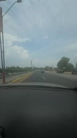 The video shows a view from the driver's seat of a car traveling down a highway on a partly cloudy day. The road has multiple lanes with some traffic. There is a sign indicating a speed limit of 60 km/h. Utility poles and trees line the side of the road. The dashboard of the car is visible in the foreground. A person speaks in the background.